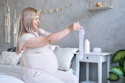 Happy pregnant woman with big belly holding baby clothes in hands at home. motherhood concept