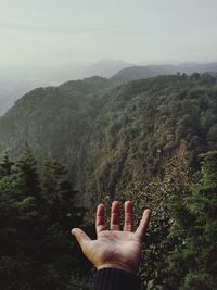 Close-up of hand against green mountain during foggy weather