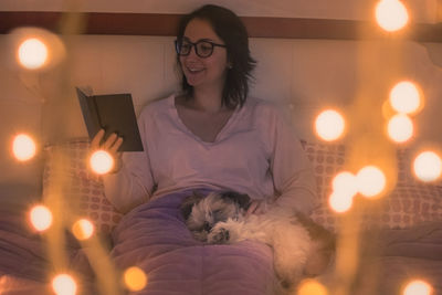 Young woman with dog reading book while sitting on bed at home