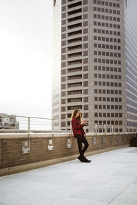 Full length of woman using smart phone while leaning on railing at terrace against building