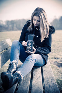 Young woman photographing while sitting on camera against sky