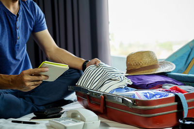 Midsection of man holding checklist and packing suitcase while sitting on bed at home