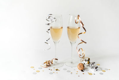 Two glasses of champagne, cork, ribbon and confetti on white backdrop.