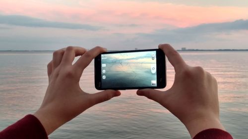 Cropped hand of person photographing lake through smart phone against sky during sunset