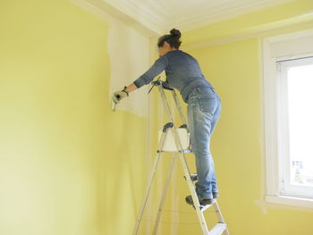 Woman on a ladder painting a yellow wall white with a paint roller