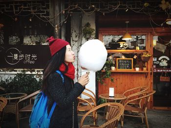 Side view of young woman holding cotton candy while standing against cafe