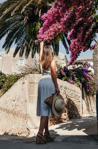 Rear view of young woman in summer dress touching beautiful pink flower in alley.