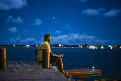 Side view of man sitting on pier by sea at night