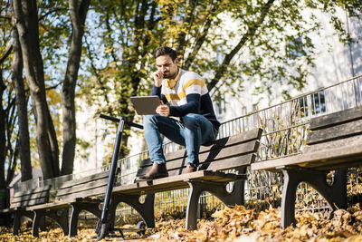 Young man using digital tablet while sitting by electric push scooter on bench at park during sunny day