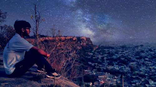 Thoughtful man sitting on cliff by cityscape against starry sky