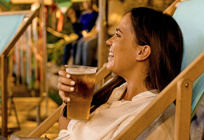 Smiling young woman drinking beer while sitting at outdoors