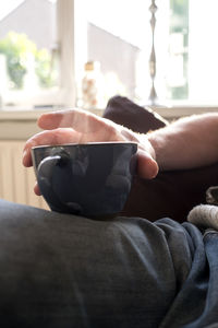 Midsection of man holding coffee while sitting on window