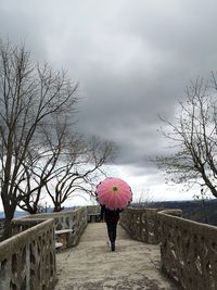 Rear view of woman with umbrella standing on footpath against cloudy sky