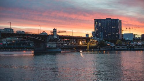 Illuminated city by river against sky during sunset