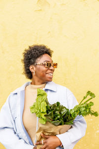 Cheerful african american female in sunglasses with paper bag of fresh green beetroot standing on yellow background wall on the street