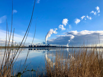 Across a lake, white smoke rises from industrial chimneys into the almost windless, blue sky