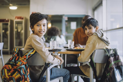 Portrait of schoolboys looking over shoulder while sitting on chair during lunch break