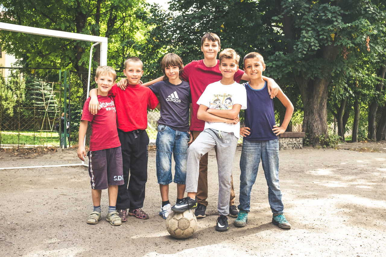 full length, looking at camera, portrait, front view, friendship, togetherness, boys, standing, child, childhood, casual clothing, day, smiling, outdoors, group of people, people, bonding, cheerful, teamwork, children only, tree, adult, sports team