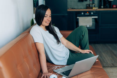 Happy young brunette sitting in the apartment on the couch chatting on the internet using a laptop.