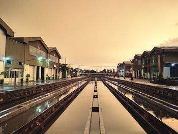 Railroad tracks by illuminated buildings against sky during sunset
