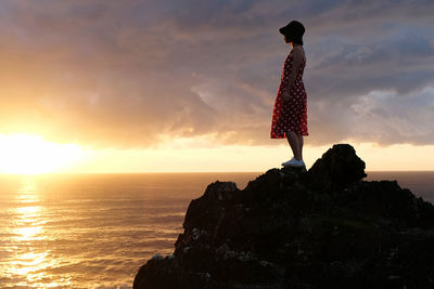 Rear view of woman standing at beach against sky during sunset