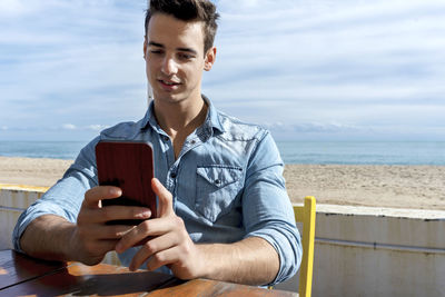 Young man using phone while sitting at cafe against beach