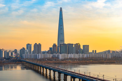 Modern buildings by river against sky during sunset