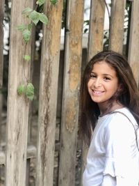 Portrait of smiling young woman standing by wooden picket fence