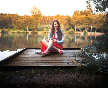 Portrait of woman sitting by pier next to lake against trees