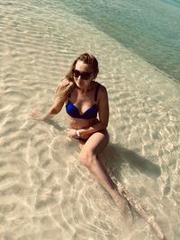 Full length of young woman wearing bikini sitting in sea on shore at beach