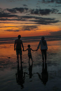 Family standing on shore at beach against orange sky during sunset