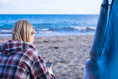 Rear view of young woman using mobile phone while sitting at beach against sky