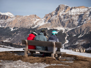 Three skiers relaxing on a wooden bench admiring the mountain panorama in front of them.