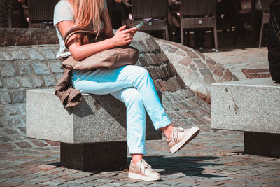 Low section of woman using mobile phone while sitting on retaining wall during sunny day