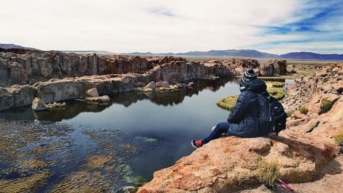 Rear view of woman sitting on rock by river against sky