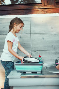 Little girl washing up the dishes pots and plates with help her sister in the outdoor kitchen