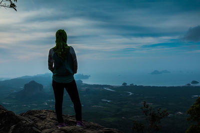 Rear view of woman standing on cliff against cloudy sky at dusk