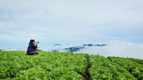 Woman photographing while crouching on agricultural field against sky