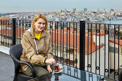 Portrait of young woman sitting by railing against cityscape