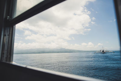 Sailboat sailing on sea against cloudy sky seen through window