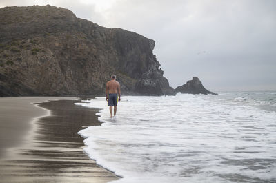 Rear view of shirtless man walking on beach with rock in cabo de gata, spain, against sky