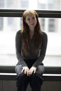 Portrait of young woman sitting on railing against window