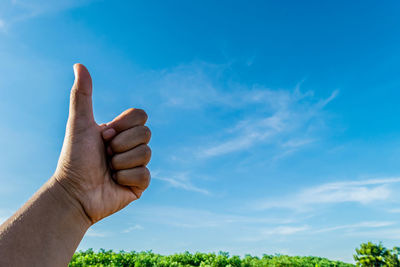 Cropped hand gesturing thumbs up on field against blue sky