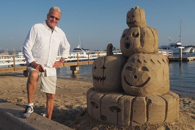 Portrait of man standing by sand sculpture at harbor against clear sky