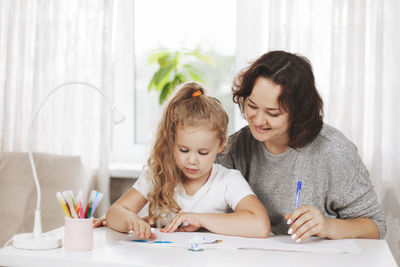 Happy mother and her smiling daughter draw on white paper, do homework for school
