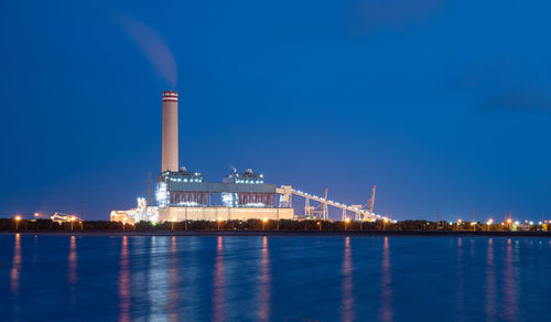 Illuminated factory by sea against clear blue sky at night