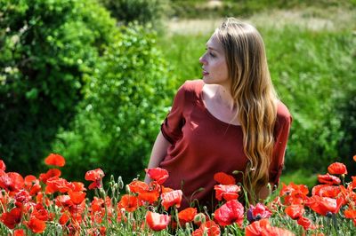 Young woman looking away while sitting on field by poppy at park