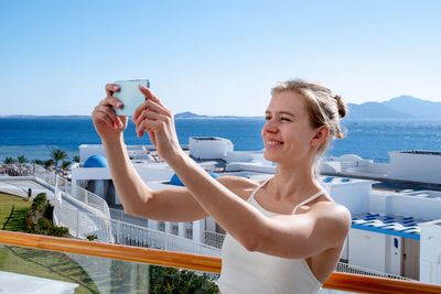 Portrait of young woman taking selfie against sea view from hotel balcony