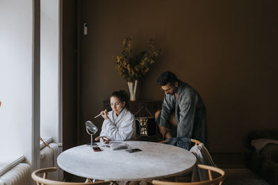 Non-binary person applying make-up while boyfriend changing clothes at home