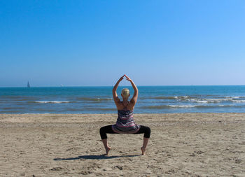 Rear view full length of woman doing yoga at beach against clear blue sky